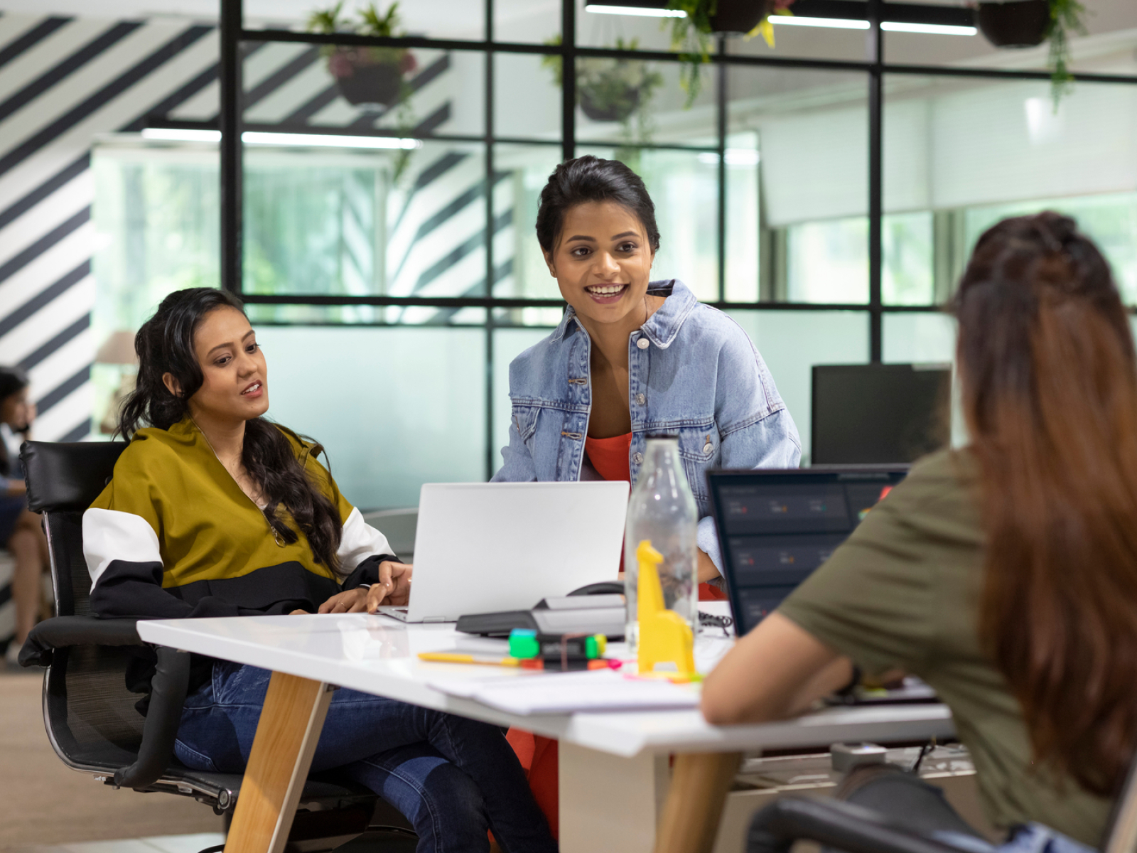 Three women are having a discussion around a table in a modern office. Laptops, bottles, and stationery are on the table. Glass walls and plants are in the background. Everyone appears engaged and smiling.