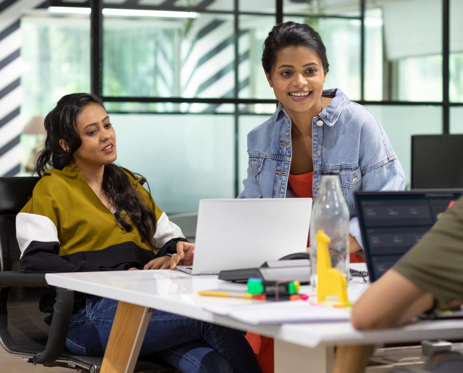 Two women collaborating in a modern office, one seated and one standing, both smiling near a laptop on a white desk with office supplies and a water bottle. Another person is partially visible in the foreground.