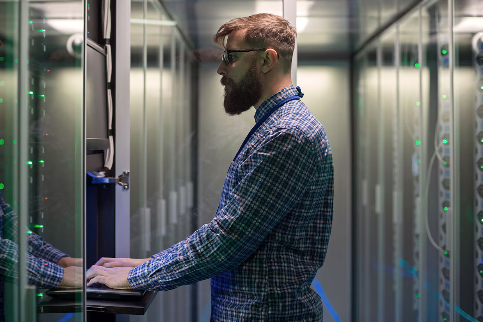 A man with a beard and glasses stands in a server room, typing on a keyboard attached to a rack-mounted monitor, surrounded by server racks with green indicator lights.