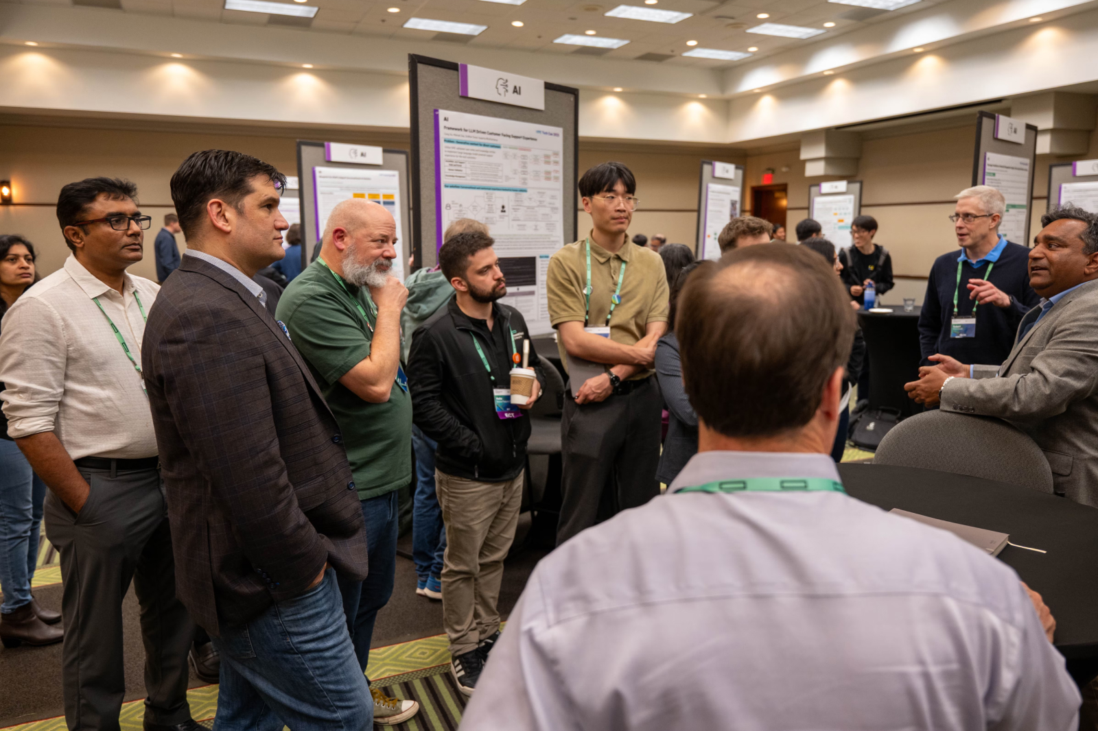 A group of people stands and listens to a speaker at a conference poster session in a large, well-lit room. Posters with scientific charts are displayed on boards in the background.