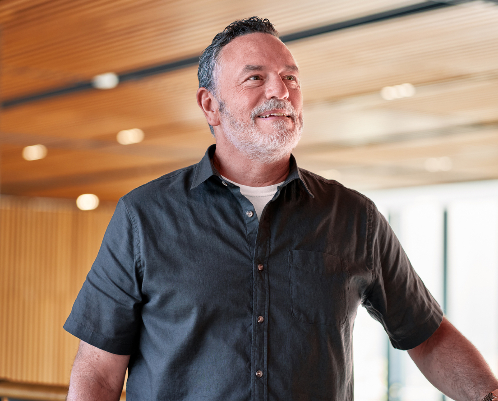 A middle-aged man with short dark hair and a gray beard smiles while standing indoors under a wooden ceiling, wearing a dark short-sleeve button-up shirt over a white t-shirt.