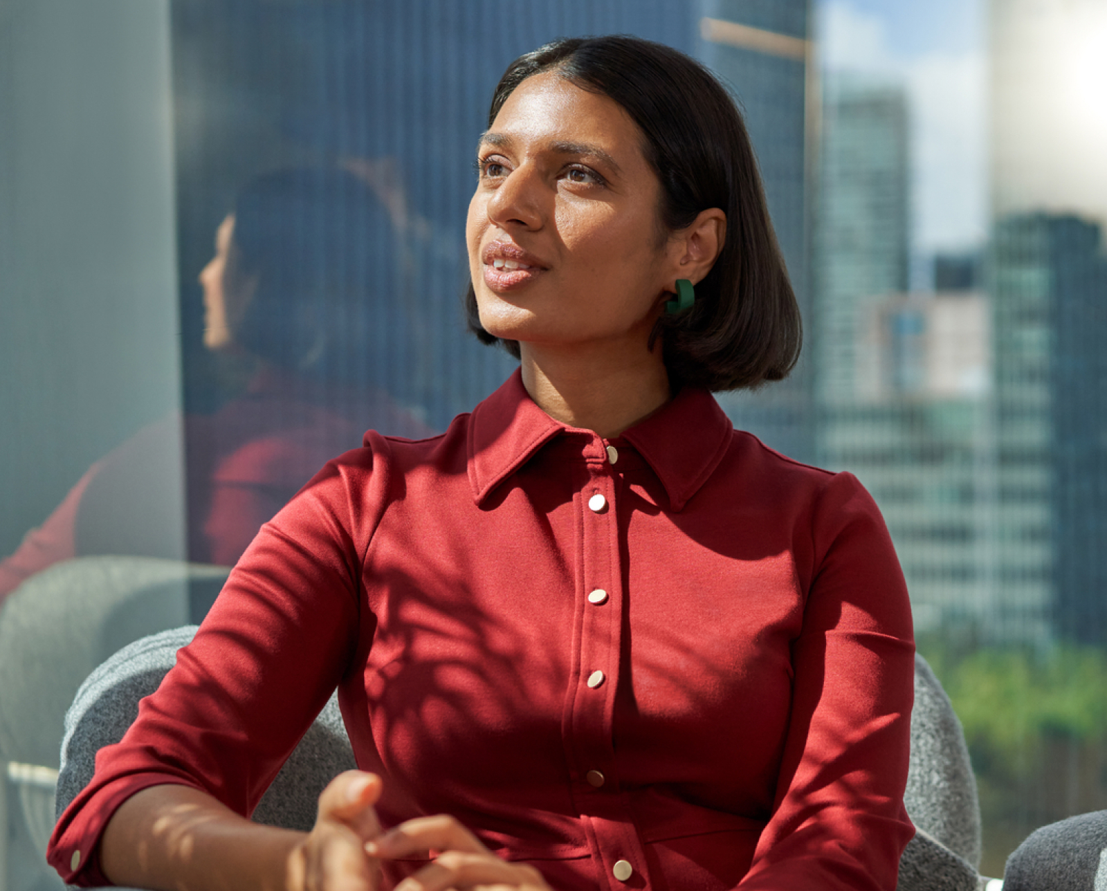 A woman with short dark hair, wearing a red button-up dress and green earrings, sits indoors on a gray chair. Sunlight casts shadows on her face and outfit. City buildings are visible through a large window behind her.