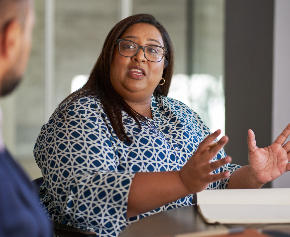 A woman with glasses and long hair, wearing a blue patterned blouse, gestures with her hands while speaking to a person across the table in an office setting. An open notebook is on the table in front of her.