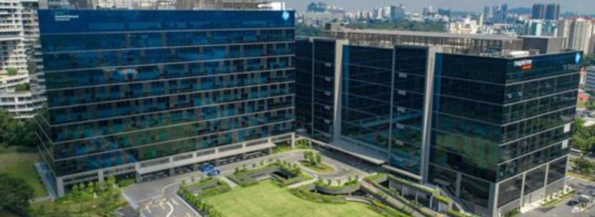 Aerial view of two modern glass office buildings with a landscaped courtyard and greenery, set against a cityscape background.