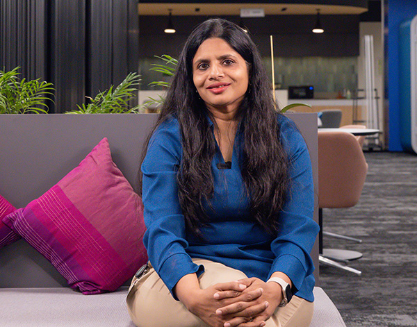 A woman with long dark hair, wearing a blue top and beige pants, sits on a gray sofa with pink cushions in a modern office lounge, smiling at the camera.