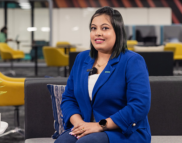A woman with straight dark hair, wearing a blue blazer and white top, sits on a gray couch in a modern office space with yellow chairs and open work areas in the background. She is smiling at the camera.