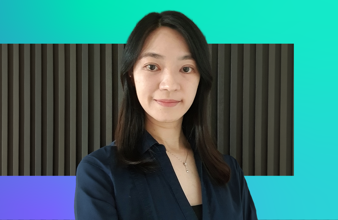 A woman with long dark hair and a navy blouse stands in front of a black ridged wall with a turquoise and purple gradient background. She is looking at the camera and smiling slightly.