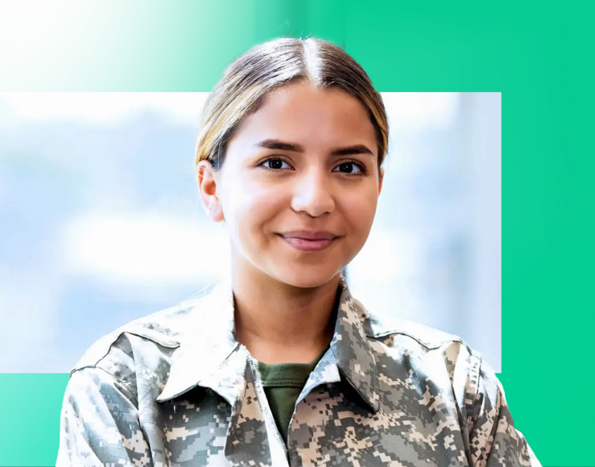 A young woman in digital camouflage military uniform smiles with arms crossed in front of a blurred blue and green background.