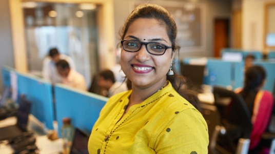 A woman wearing glasses and a yellow top smiles at the camera in a modern office, with coworkers working at desks in the background.