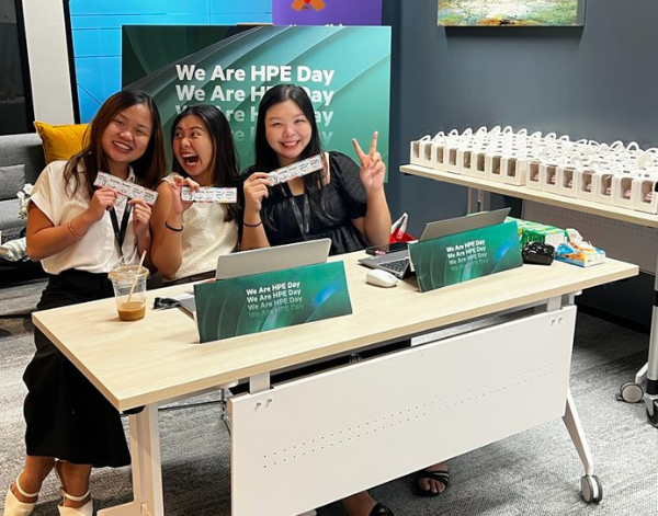 Three women sit at a desk, smiling and posing with lanyards. Behind them is a sign that reads “We Are HPE Day,” and a table with rows of small white boxes. Laptops and drinks are on the desk.