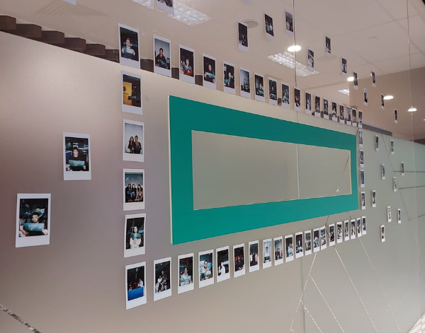 A glass wall displays numerous instant photos arranged around a large green rectangular panel and a mirror, showcasing various people in small groups or alone, creating a collage-like decoration in an office setting.