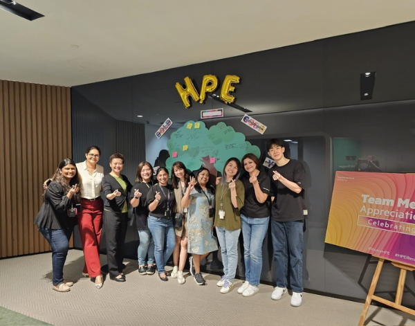 A group of nine people smiling and posing with finger hearts in front of a wall decorated with gold "HPE" balloons and colorful notes. A "Team Member Appreciation" sign is visible on the right.