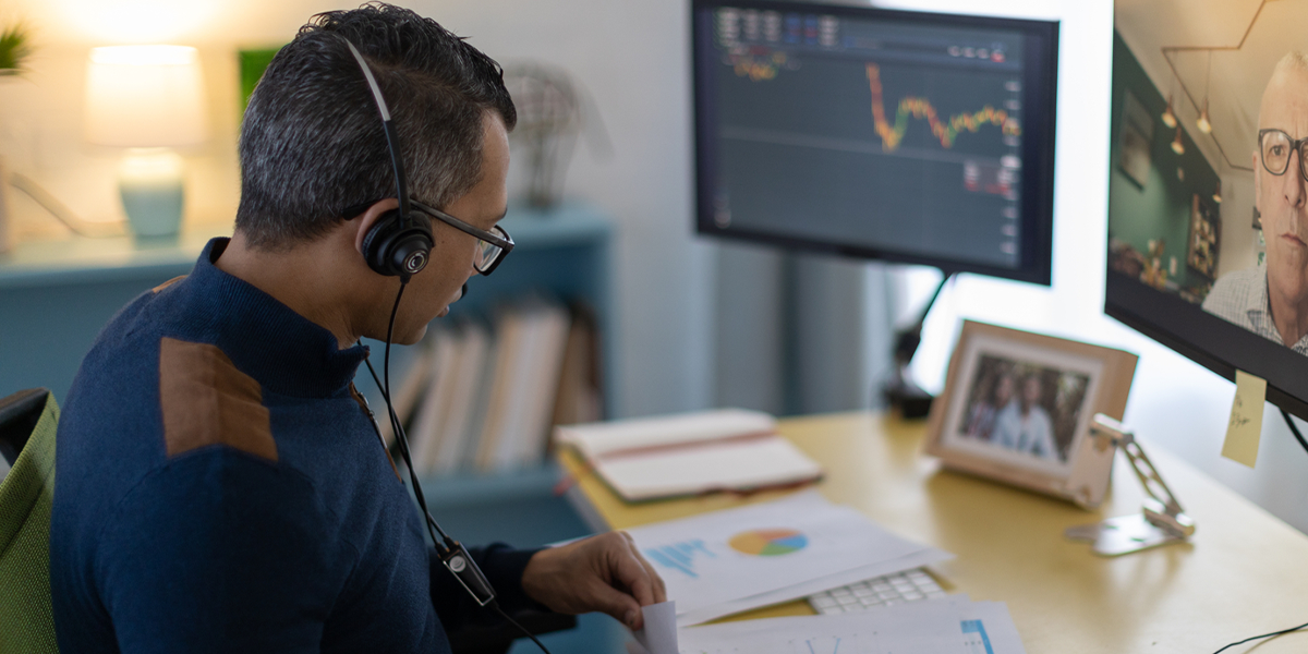 A man wearing a headset sits at a desk with papers, looking at computer monitors showing a video call and a stock chart. A framed family photo and office supplies are on the desk.