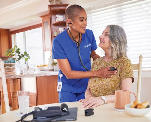 Nurse listening to the heart of a smiling, senior woman who is sitting at a kitchen table.