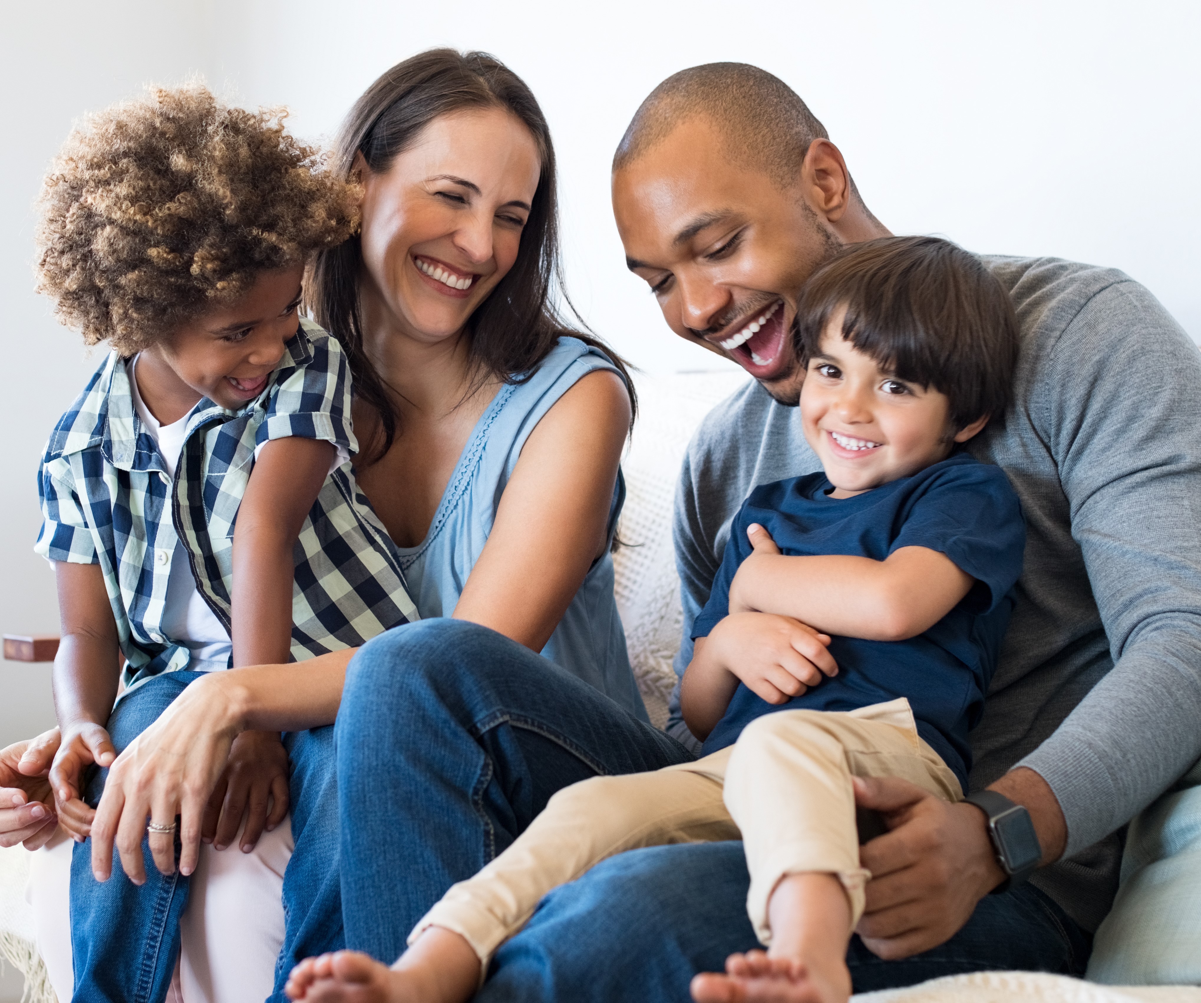 A cheerful family of four sits together on a cozy couch smiling and laughing.