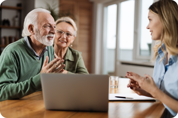 A social worker sits at a table with a senior couple in their home sharing information with them on her laptop. 