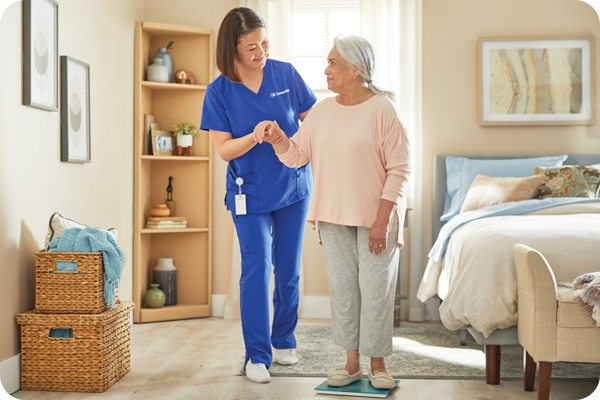 A clinician standing next to and holding the hand of a woman as she walks in her bedroom.