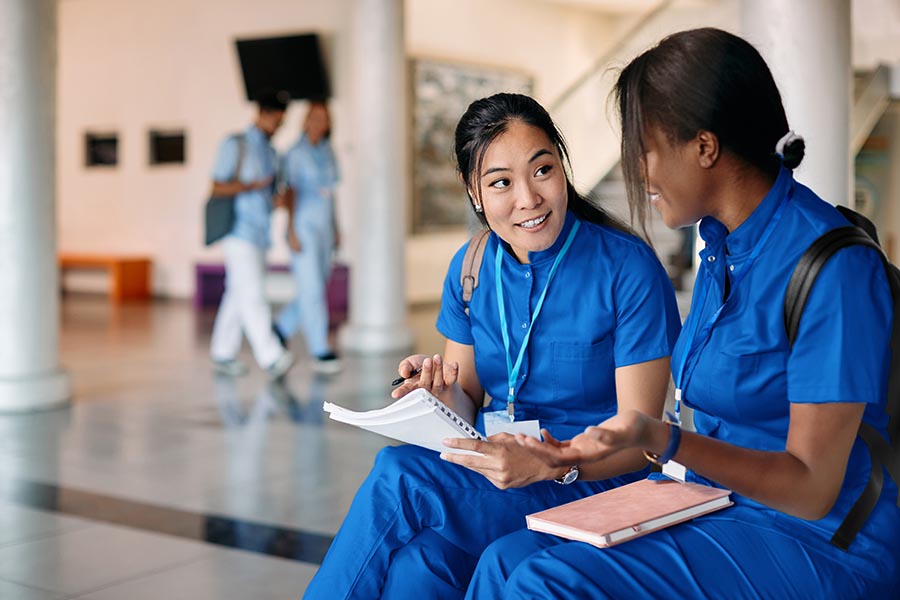 Two female clinical students wearing backpacks and blue scrubs sitting in foyer with notebooks in conversation
