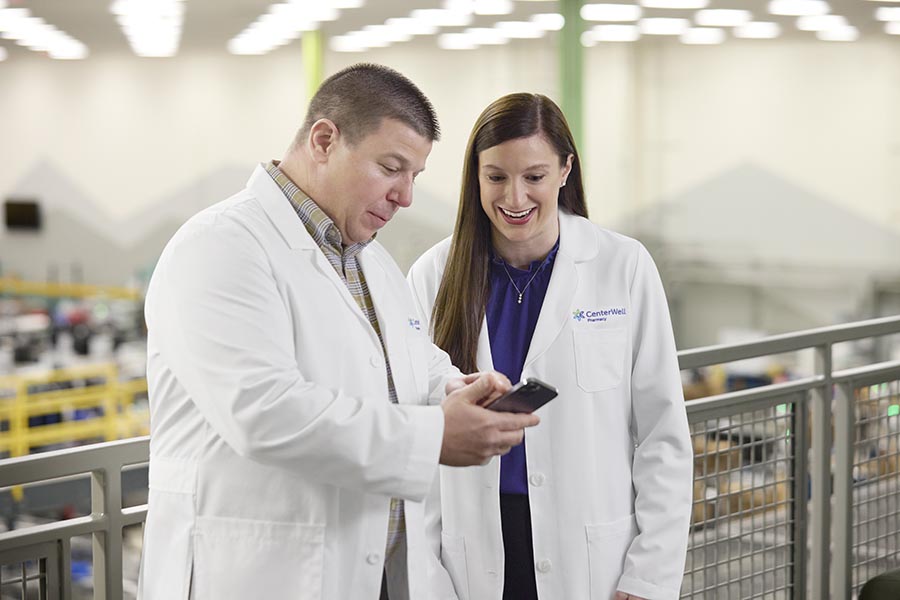 Male pharmacist in white lab coat showing a female pharmacist something on his tablet in a pharmacy setting