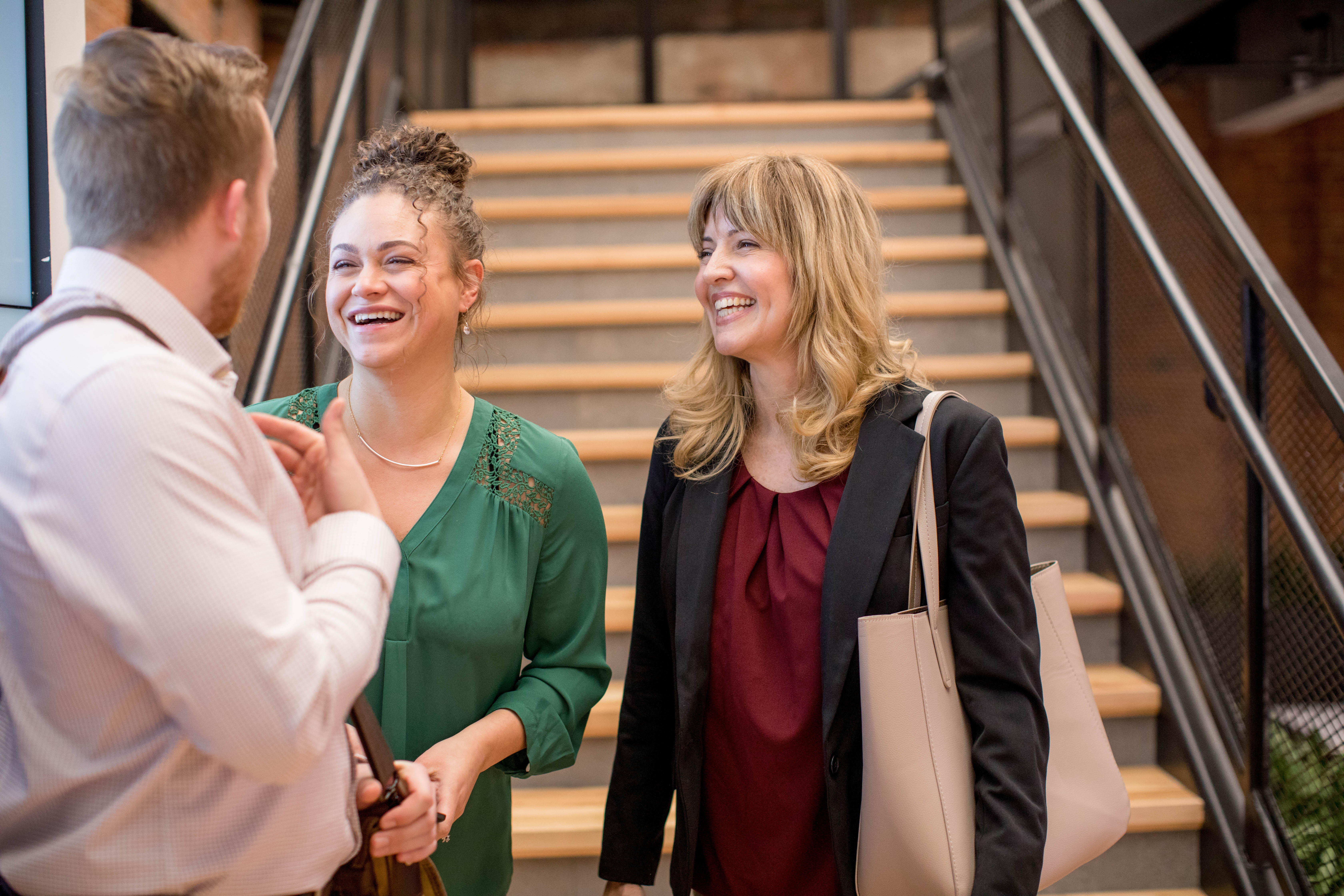 Three professionals standing at the bottom of an office stairwell sharing a laugh together.