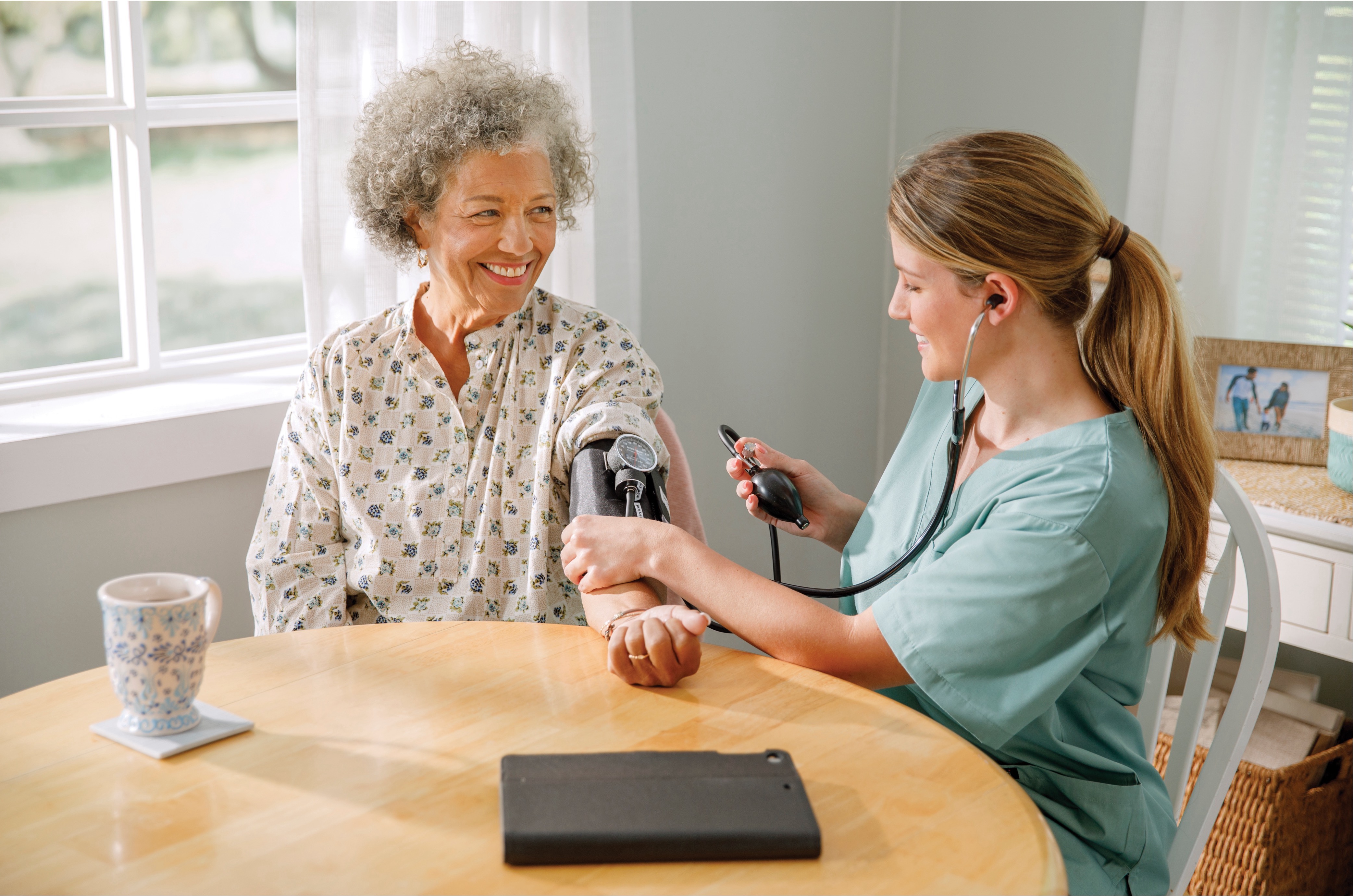 Female nurse taking blood pressure of an elderly woman while seated at a table in a home setting.