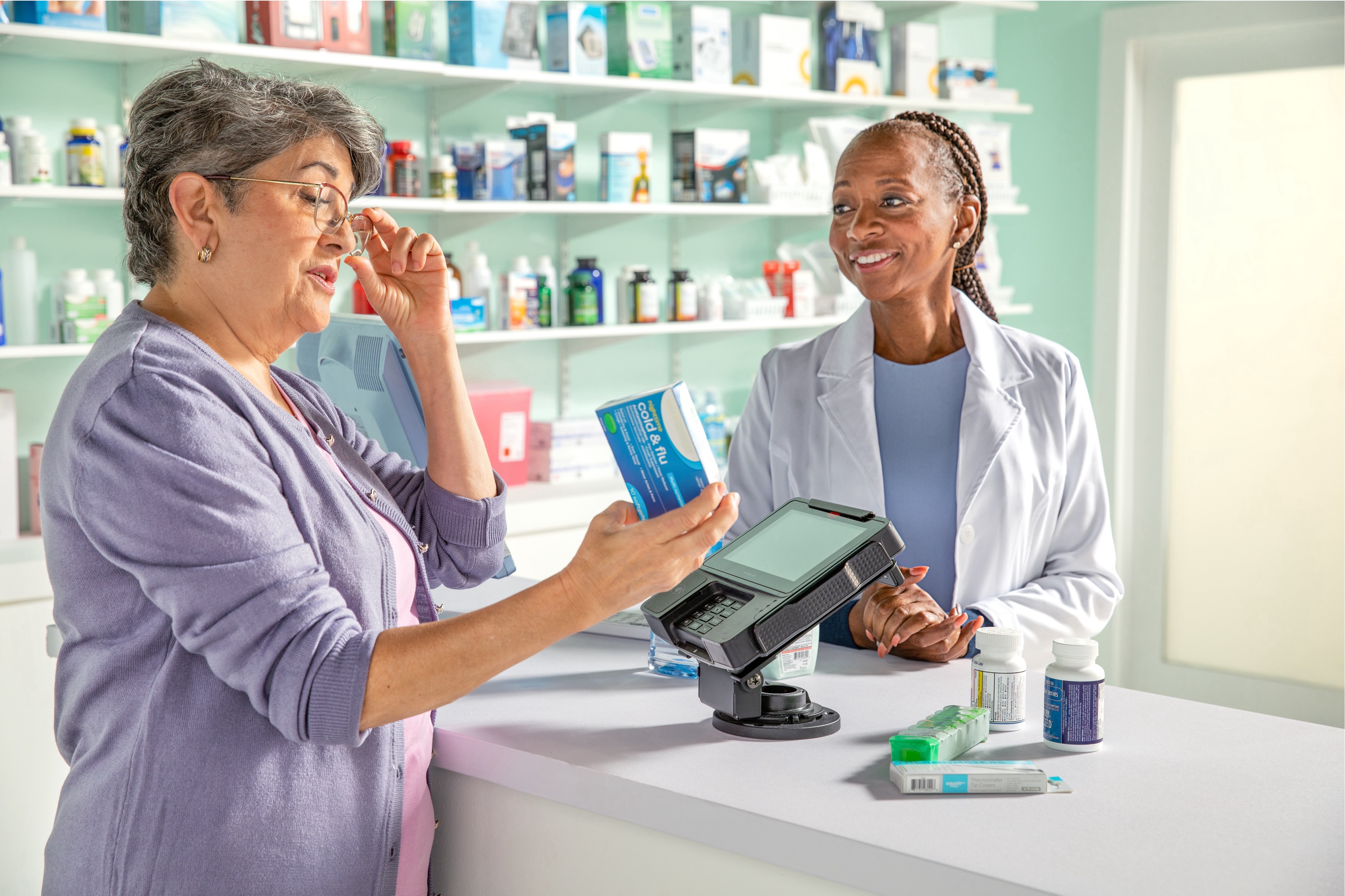 Female pharmacist smiling behind the counter at a pharmacy while an elderly woman closely examines medication.