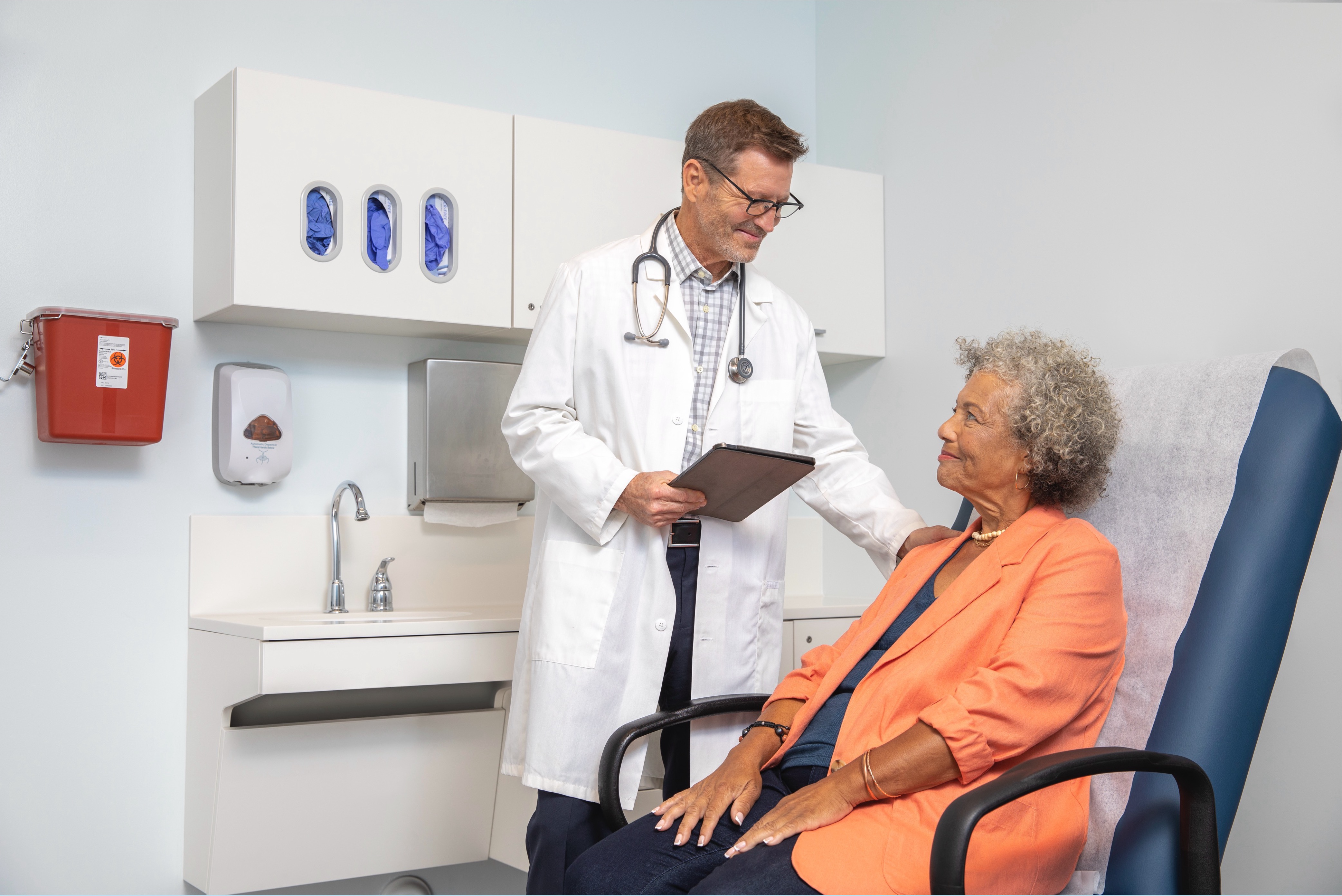 Male physician holding a clipboard consulting with a female patient in an exam room.