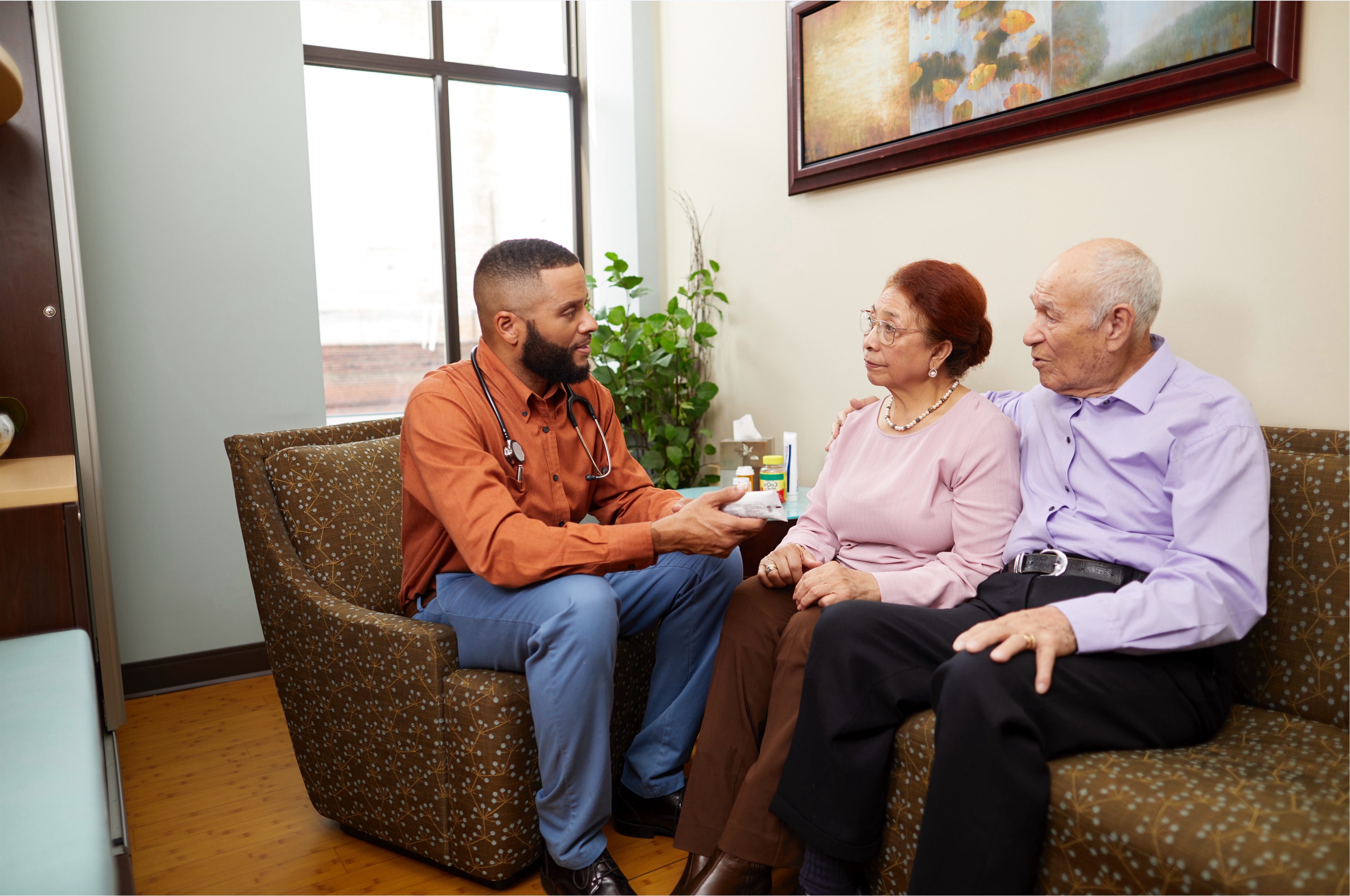 Male social worker speaking with an elderly couple while seated on couches in a home environment.