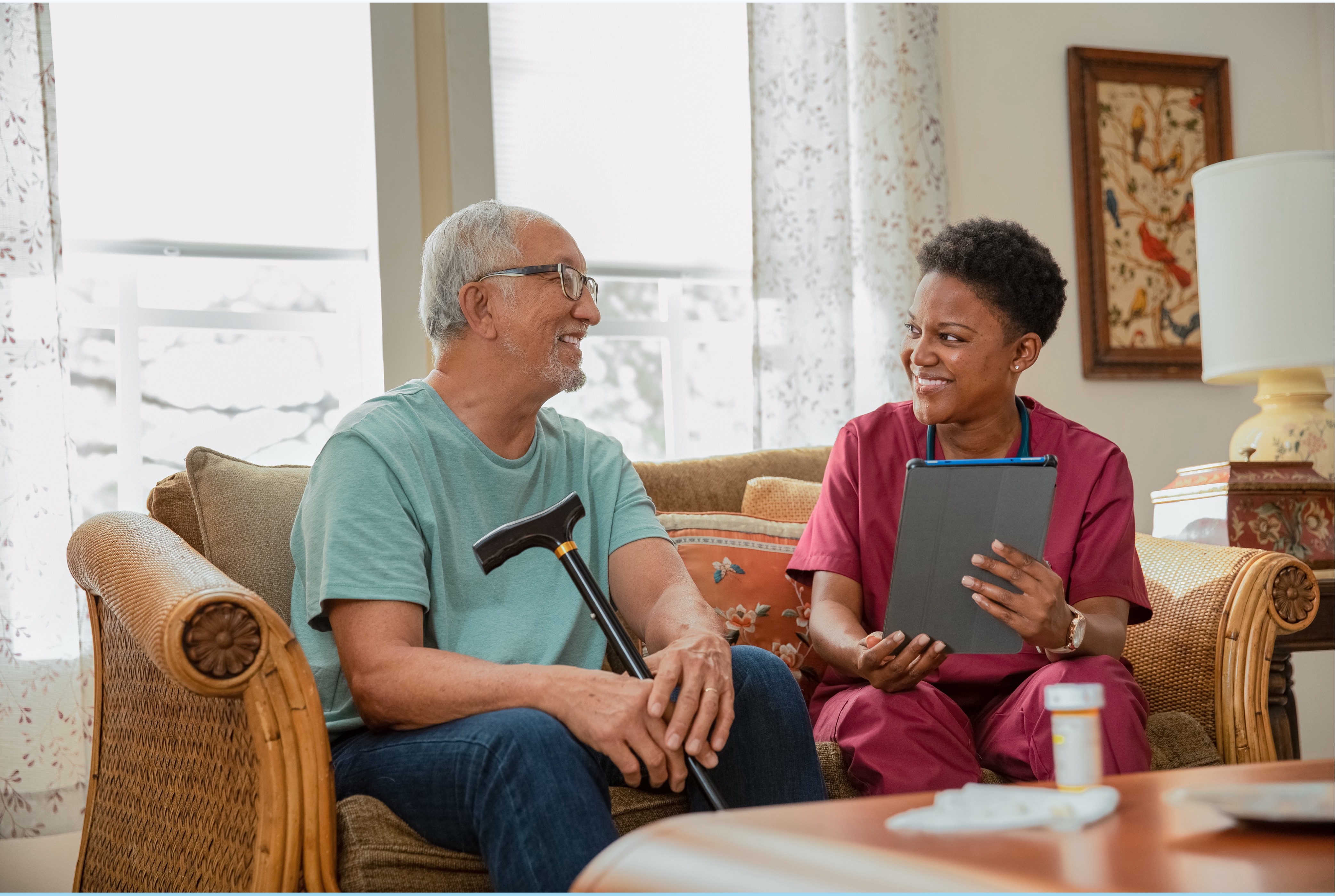 Female wearing scrubs holding a clipboard talking to an elderly man with a cane seated on a couch in a home.