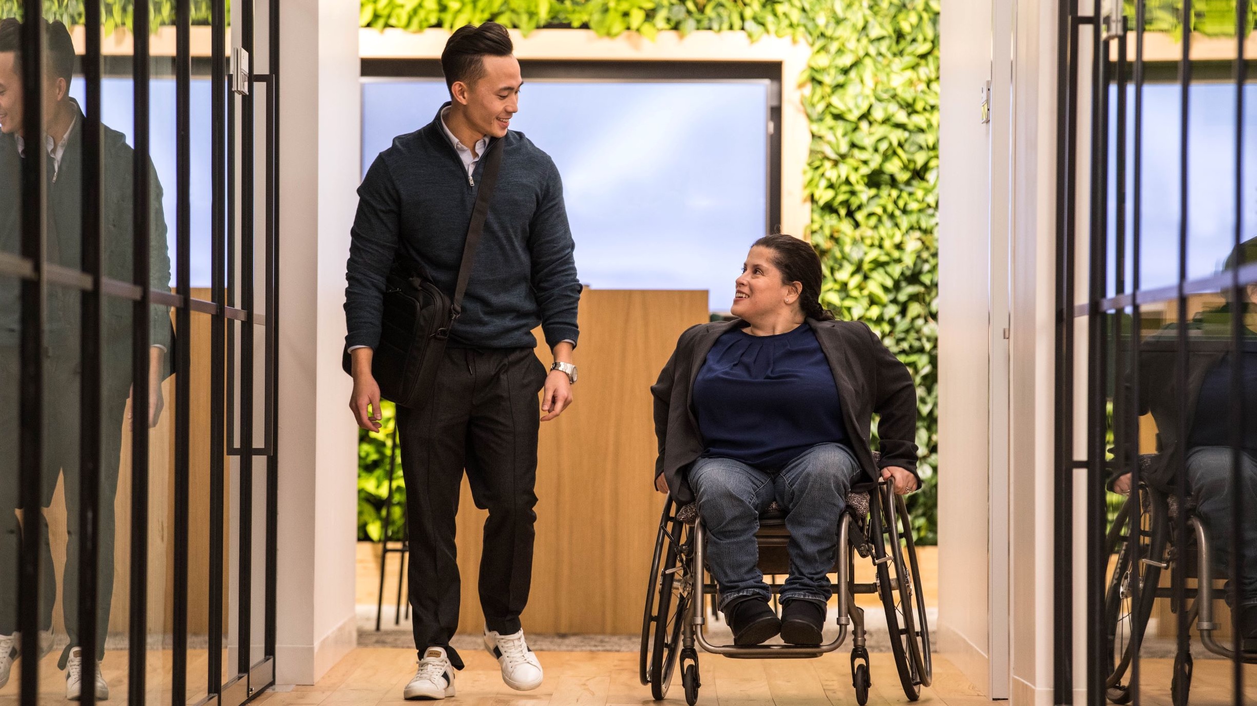 Young male walking with women in wheelchair down an office hallway. 