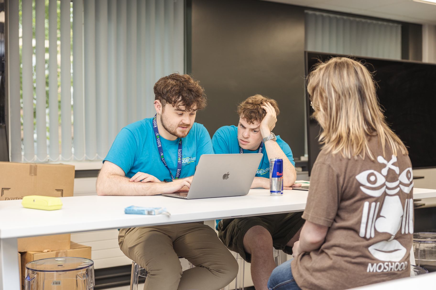 Two young men in blue t shirts look at laptop enthusiastically, with a young person presenting 