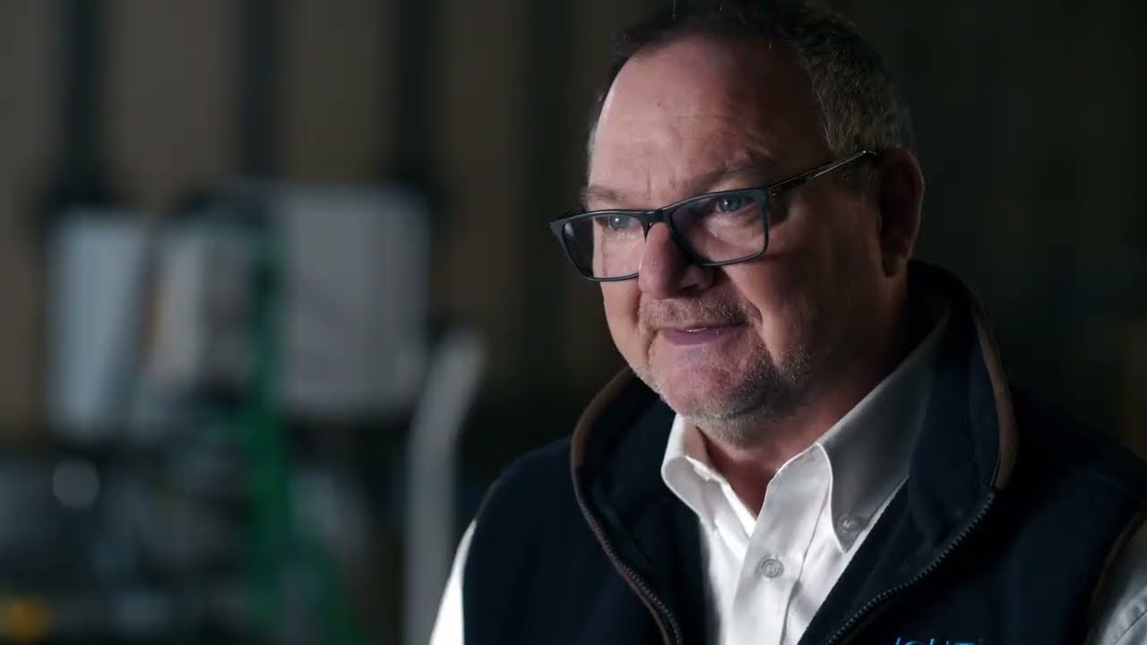 Portrait of an adult man wearing glasses and business-casual clothing, smiling slightly in a softly lit workshop or industrial environment.