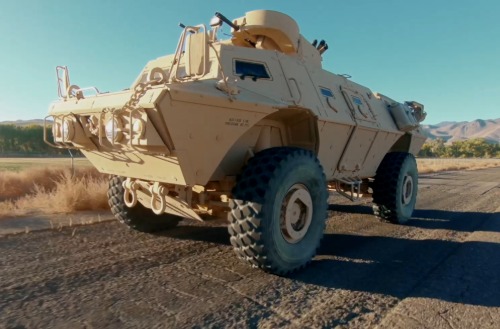 Textron Systems armored tactical vehicle driving on a paved road in a rural landscape with mountains in the background.