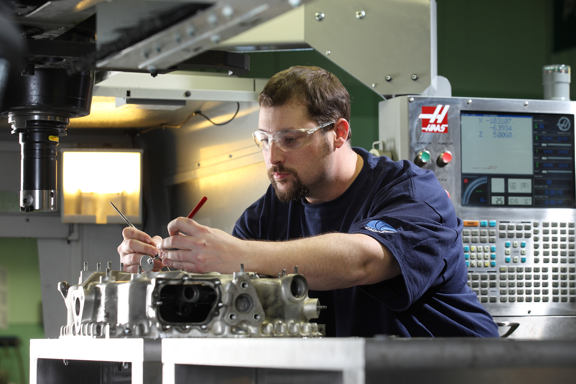 Cadorath machinist in safety glasses carefully measuring a precision-machined metal component on a CNC machine in a Louisiana manufacturing facility.