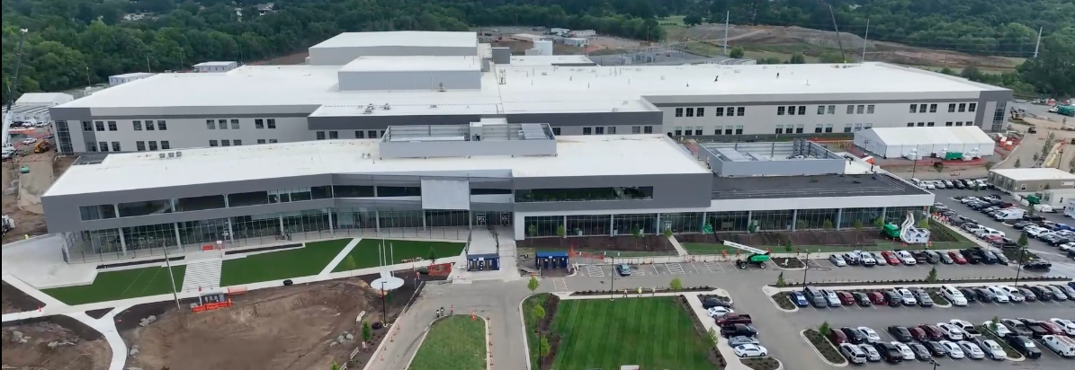 An arial view of the Concord manufacturing plant, the plant is primarily gray with green full trees in the background. The parking lot is also visible with many cars and there shows some areas still being built on with brown dirt