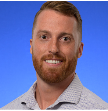 Headshot image with blue backdrop of Jake Monroe, a Caucasian male with a red beard, smiling