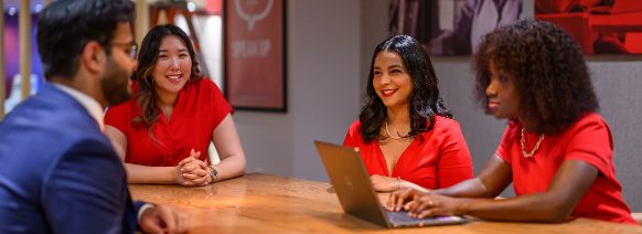 a group of four diverse individuals are in a meeting at a wooden conference table. The three women are in red professional dresses and one male in a navy suit. They are smiling.