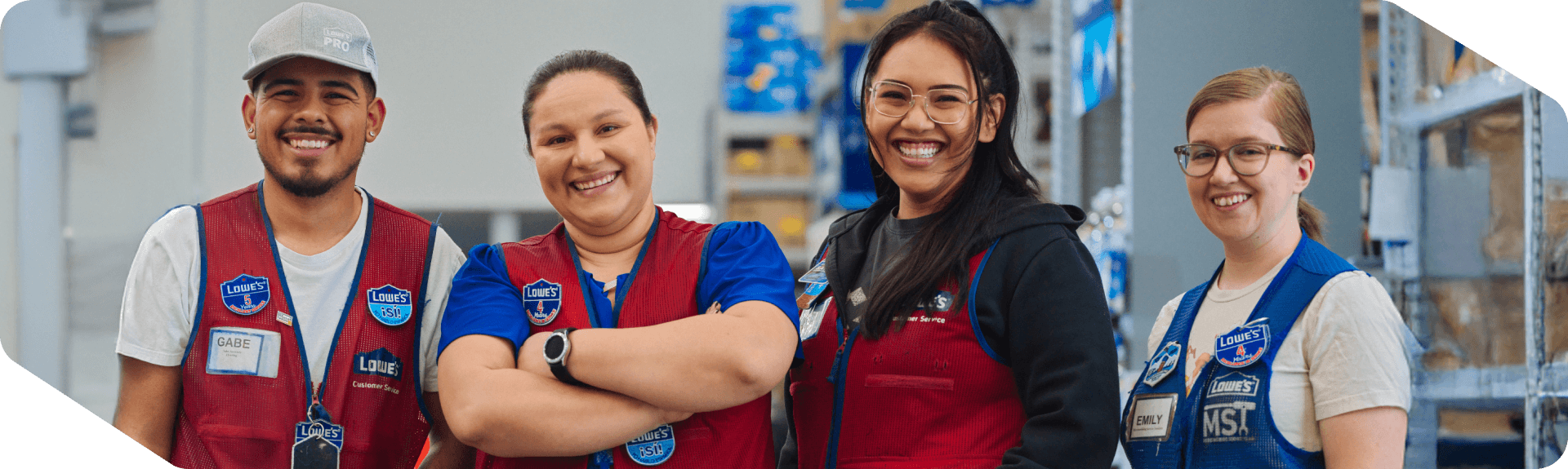 Four happy Lowe's associates in red and blue vests standing together in a store aisle.