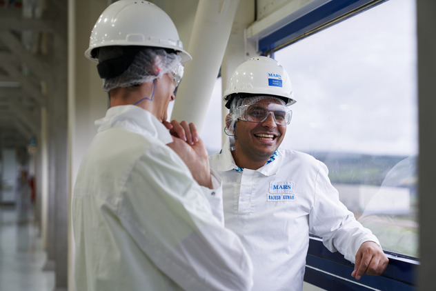 An image of 2 Mars Associates dressed in white Mars uniform whilst wearing protective hard hats with hair nets underneath and protective goggles engaged in conversation and laughing.