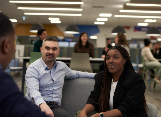 Image of two Associates sitting on a sofa, speaking to another Associate. 