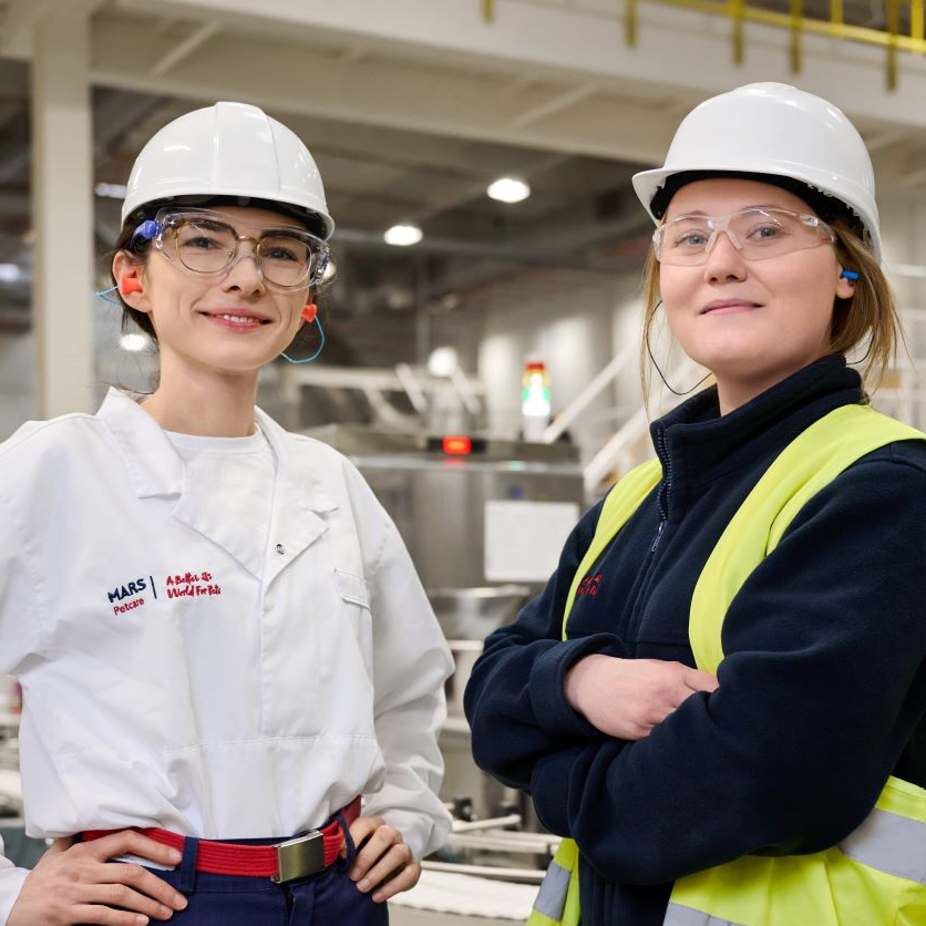 An image of 2 Mars Associates wearing protective hard hats and protective goggles, the associate on the left is wearing a lab coat tucked into their jeans with their hands on their hips and the associate on the right is wearing a navy fleece and a hi visibility vest with their arms crossed. The associates are standing side by side and smiling. 