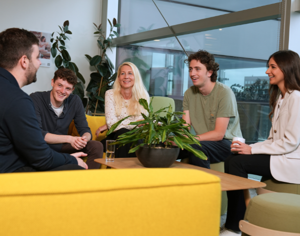 A group of colleagues gathered around a table, smiling and engaging in discussion in an office with natural light and plants.
