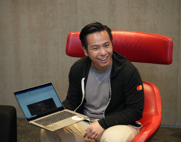 A man sitting in a red chair, smiling while working on his laptop in a modern office setting.