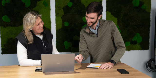 Two colleagues smiling while working together with one colleague pointing to the laptop on the wooden bench