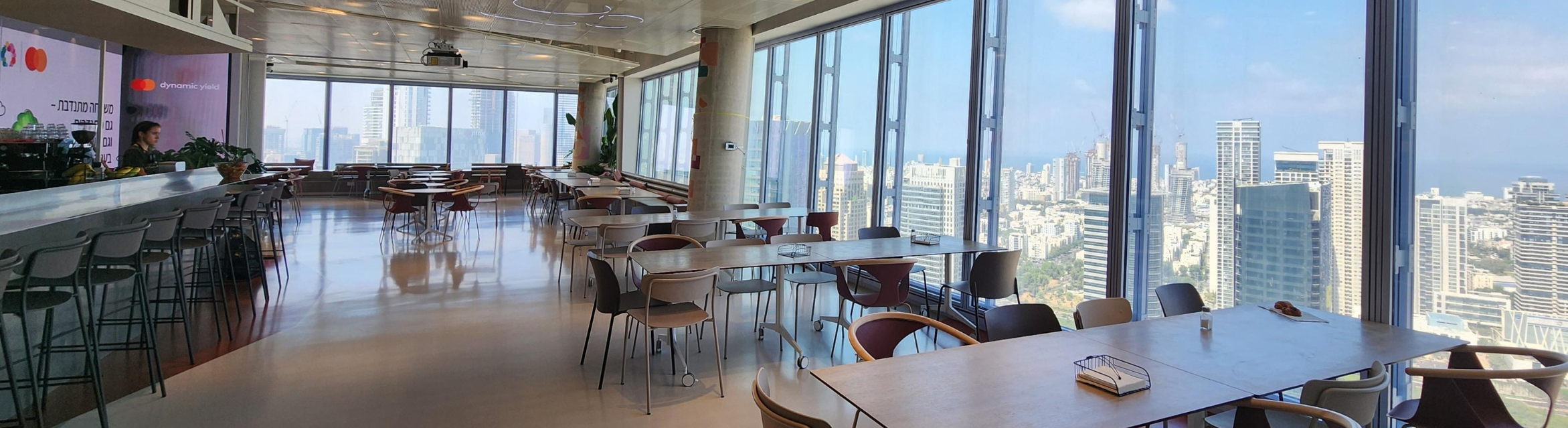 The canteen area at Mastercard's Tel Aviv office, featuring tables and chairs, with expansive floor-to-ceiling windows offering panoramic views of the city skyline.