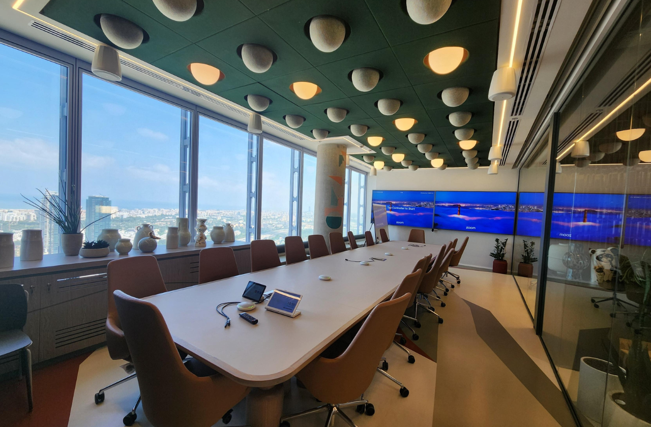 A spacious conference room in Mastercard's Tel Aviv office, featuring a large table surrounded by plenty of chairs. The room is equipped with large TV screens, unique ceiling lighting, and offers views of the city through expansive windows.