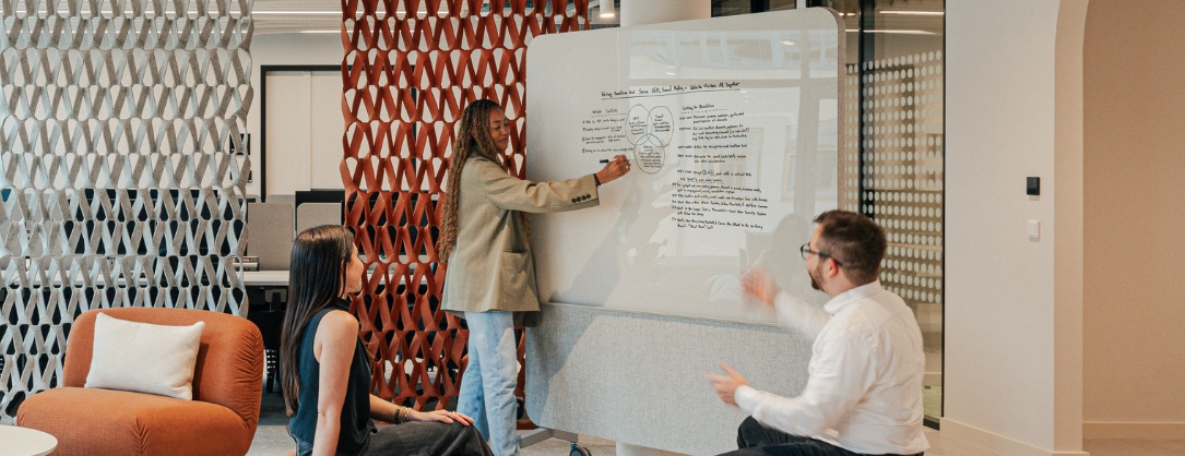 a woman standing and pointing at a whiteboard with text, while man and woman sitting down are listening