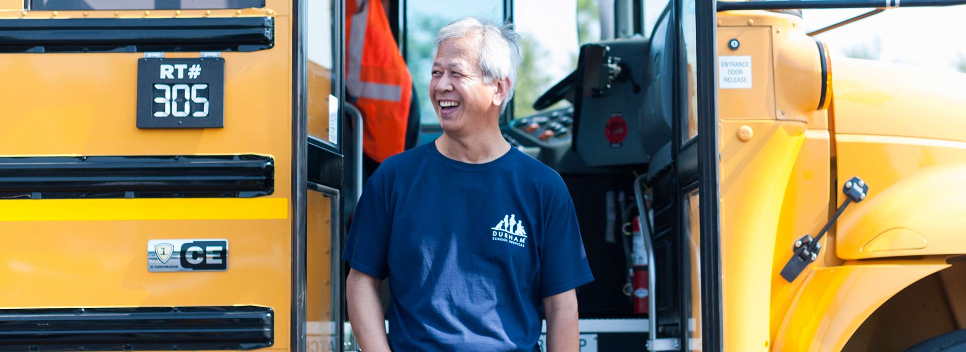 A cheerful male school bus driver stands by the front door of the bus, wearing a big smile