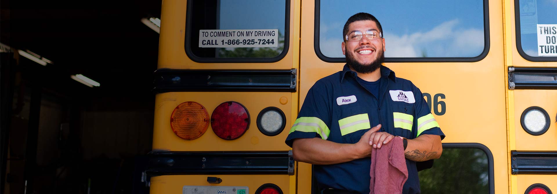 A school bus mechanic wearing a smile and standing confidently in front of a school bus, showcasing pride in their work.