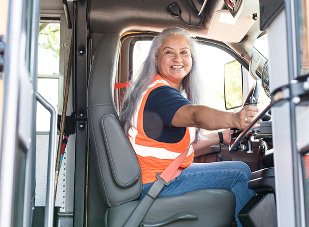 A smiling female school bus driver seated behind the wheel, attentively observing the area through the open bus door